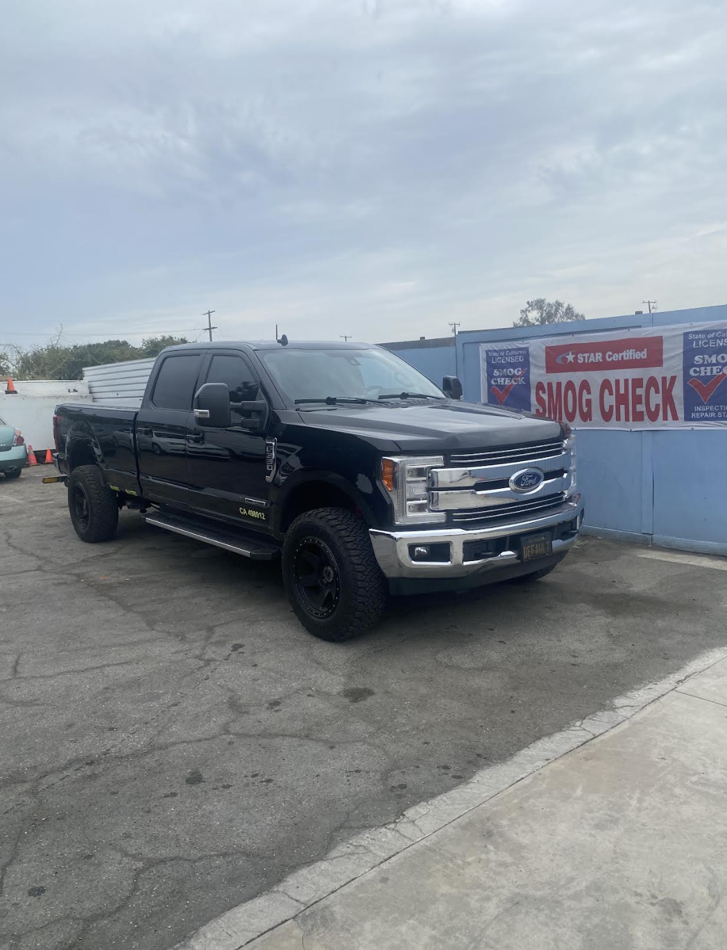 Black Ford Super Duty pickup truck parked by the STAR Certified SMOG CHECK banner